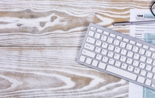 Overhead view of tax form, pen, keyboard, and reading glasses on fading white desktop.